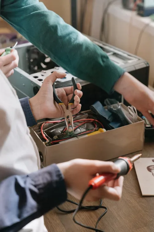 Hands use pliers and a soldering iron to work on an open electronic device.